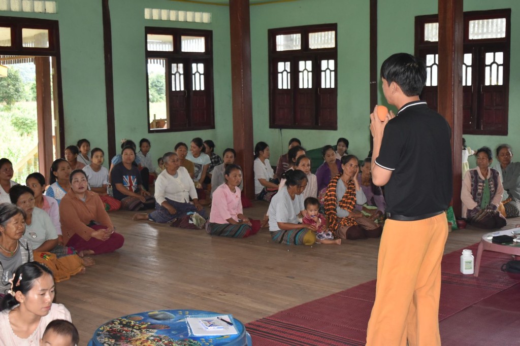A volunteer speaking to women and children seated on the floor of a village hall